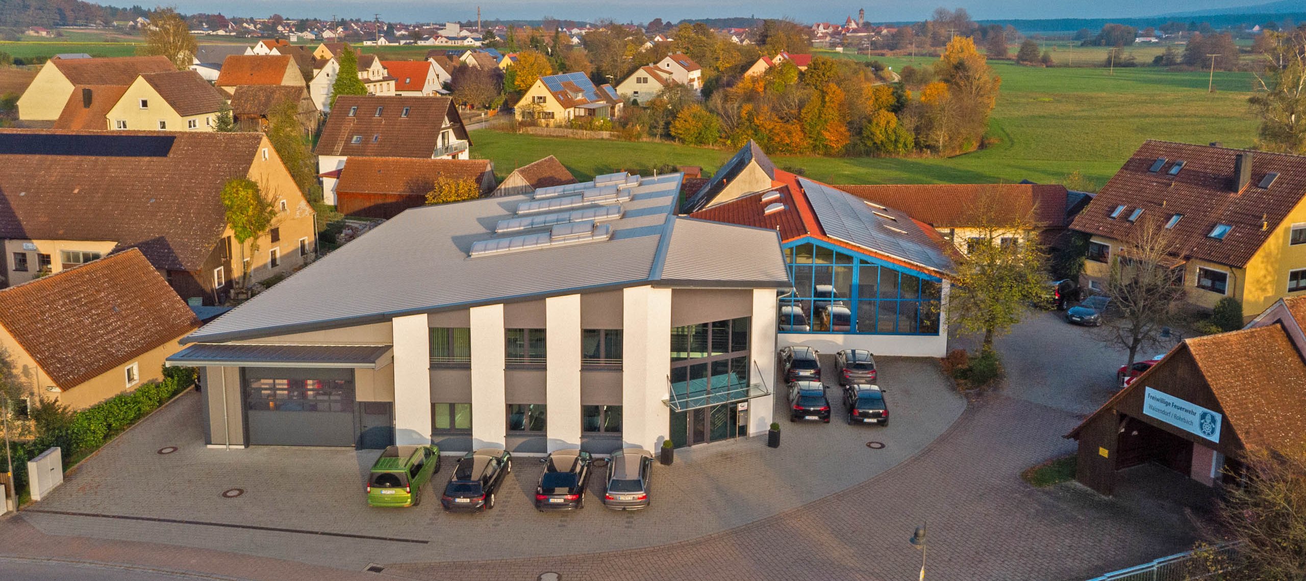 Firmengebäude der WU Mechanische Werkstätte Blick auf das Firmengebäude der WU Mechanische Werkstätte GmbH mit mehreren geparkten Autos davor, umgeben von Wohnhäusern und grüner Landschaft.